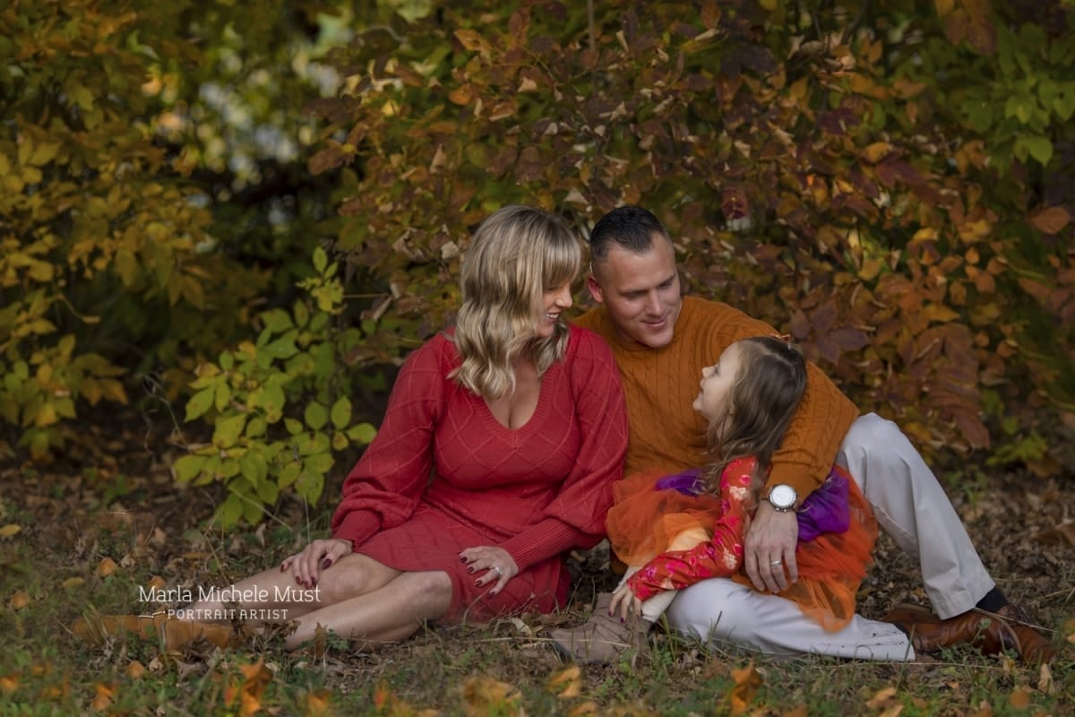 Family portrait of a mother, father and daughter dressed in red and orange, sitting on the forest floor and smiling at each other | Detroit, Michigan