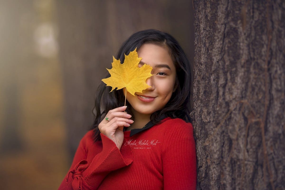 A high school senior poses for her portrait by holding a golden leaf to conceal on of her eyes as she smiles at the camera in a Detroit-area forest