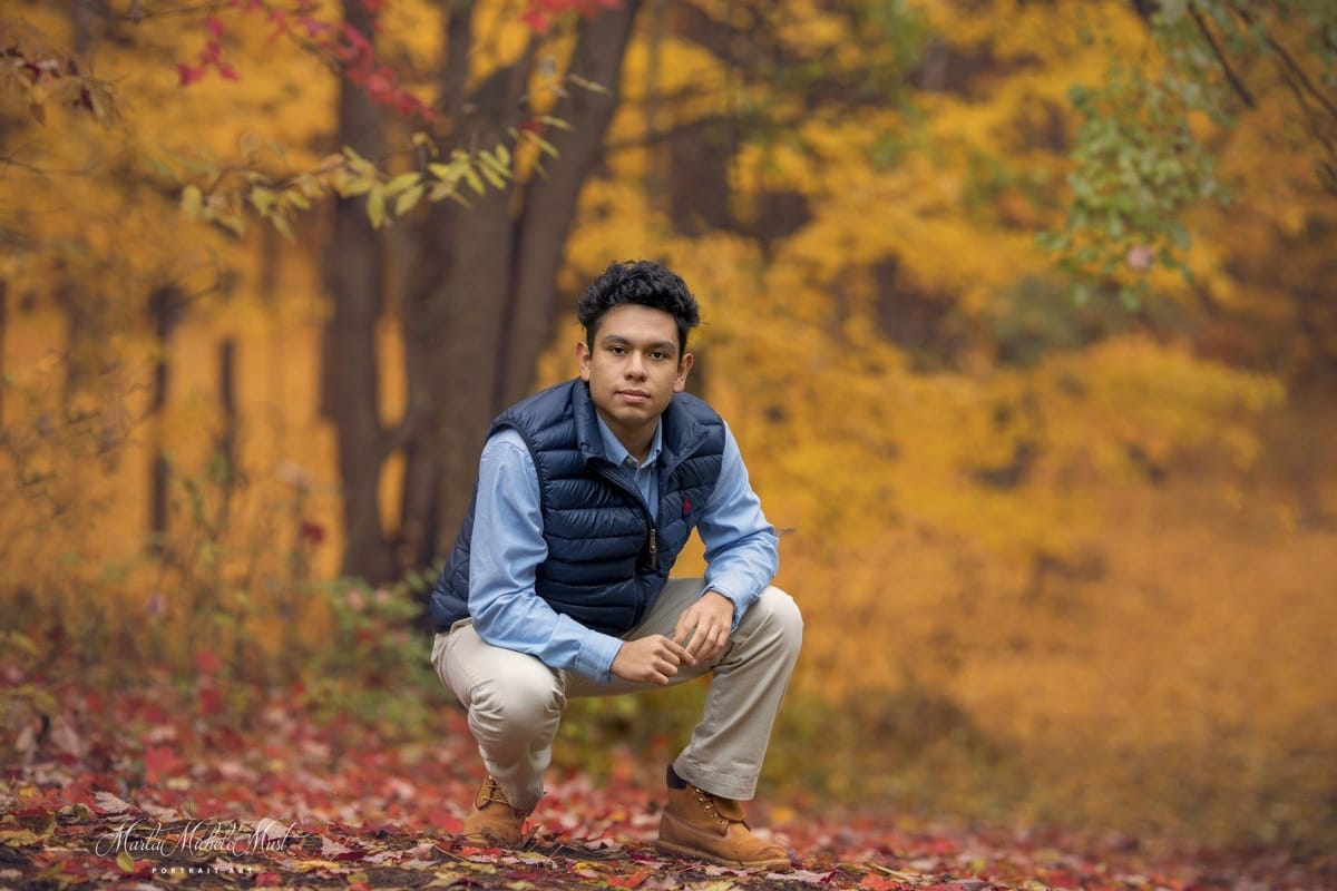 A senior portrait of a young man crouched among golden leaves in a a Detroit forest, beautifully captured during a senior graduation photoshoot photographer.