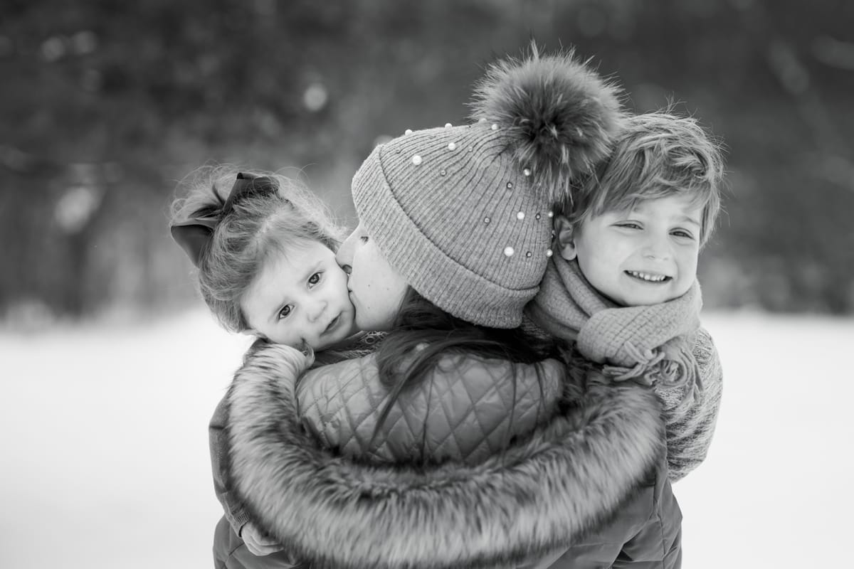 A winter family photoshoot in black and white. A woman has her back to the camera holding her two children while snow falls in Michigan