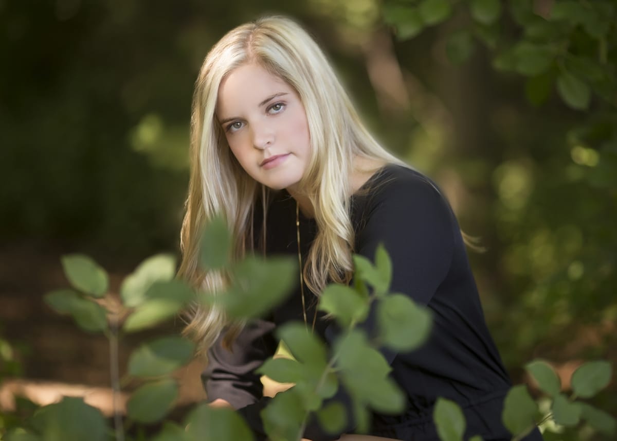 A young woman with blonde hair and a black long sleeve shirt poses among green leaves and branches in a Michigan forest.