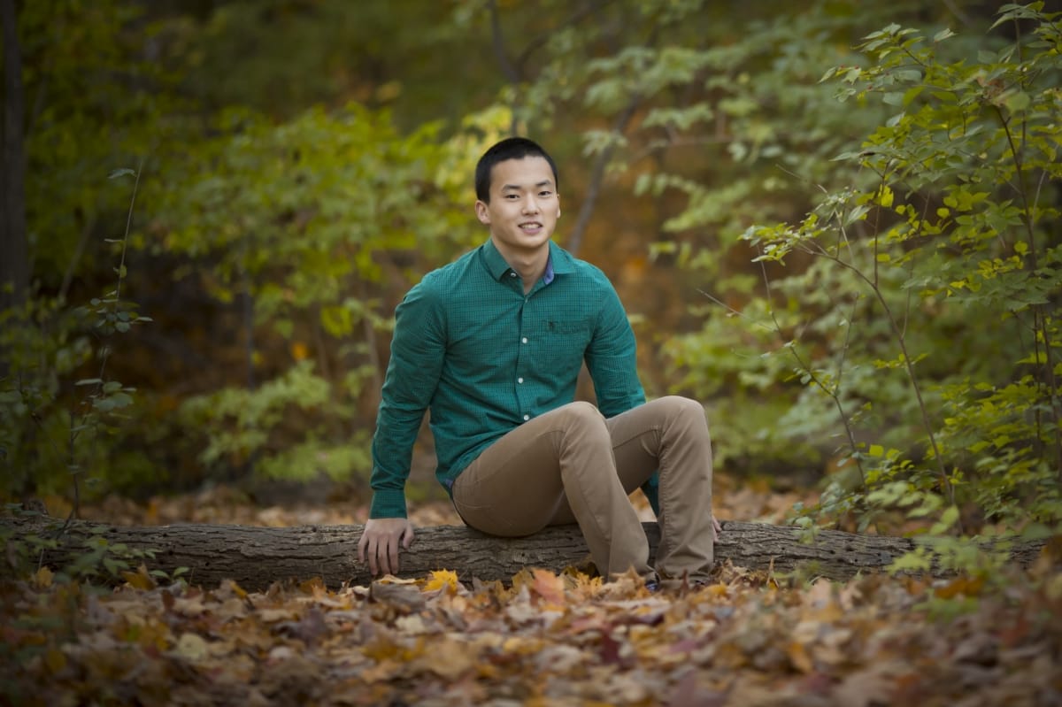 In this high schools senior portrait, a young man sits on a log and smiles for his senior portrait in a Detroit-area forest.