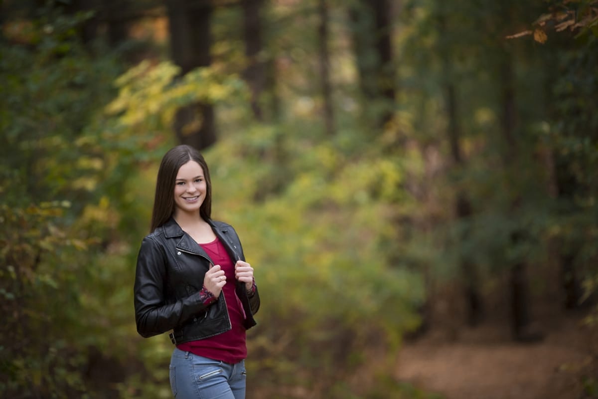 A cool and grunge-inspired photoshoot of a senior holding the lapels of her leather jacket as she poses in a Detroit forest.