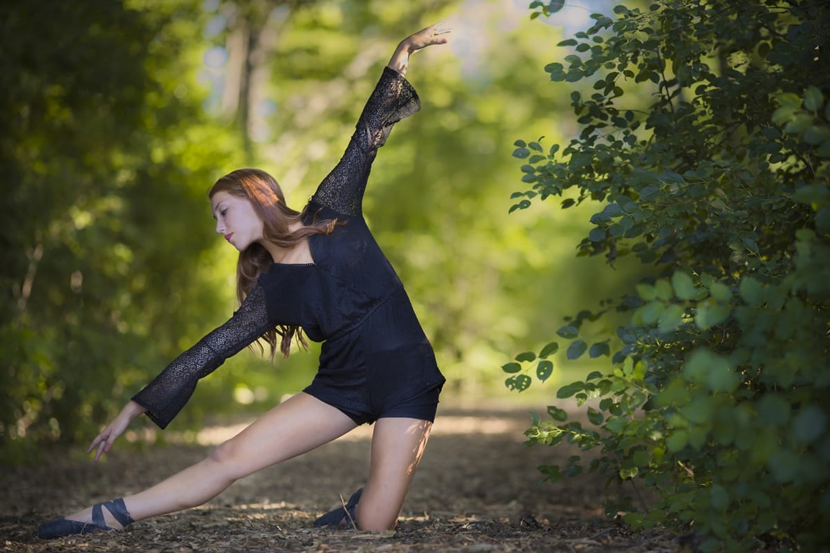 Dancer gracefully embodies a lyrical pose, gracefully extending her arms long and bending towards her legs, captured by a talented Detroit photographer in a Michigan forest.