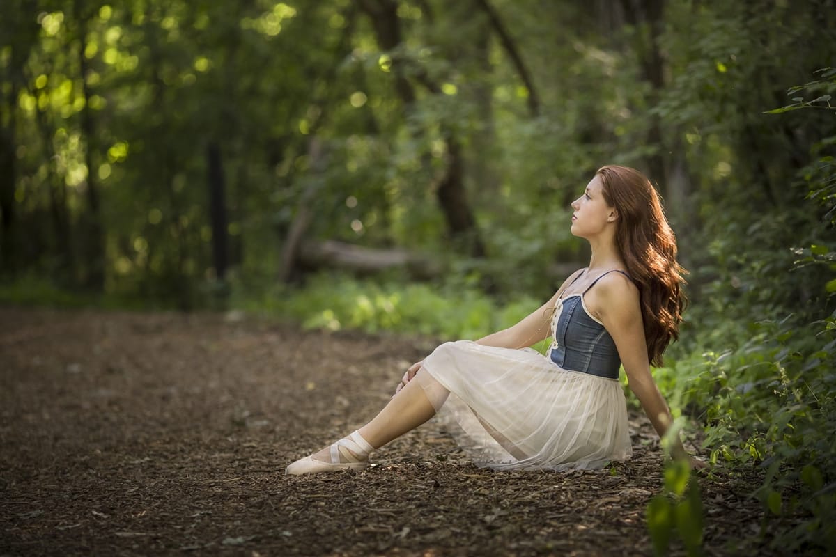 Dancer sits peacefully in a forest clearing while wearing pointe shoes, captured by a Detroit photographer during a dance photoshoot.