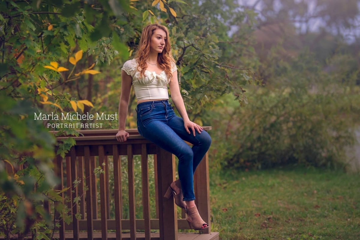 An outdoor senior high school graduate photoshoot depicts a young woman in a Detroit-area forest sitting on a fence and gazing onward.