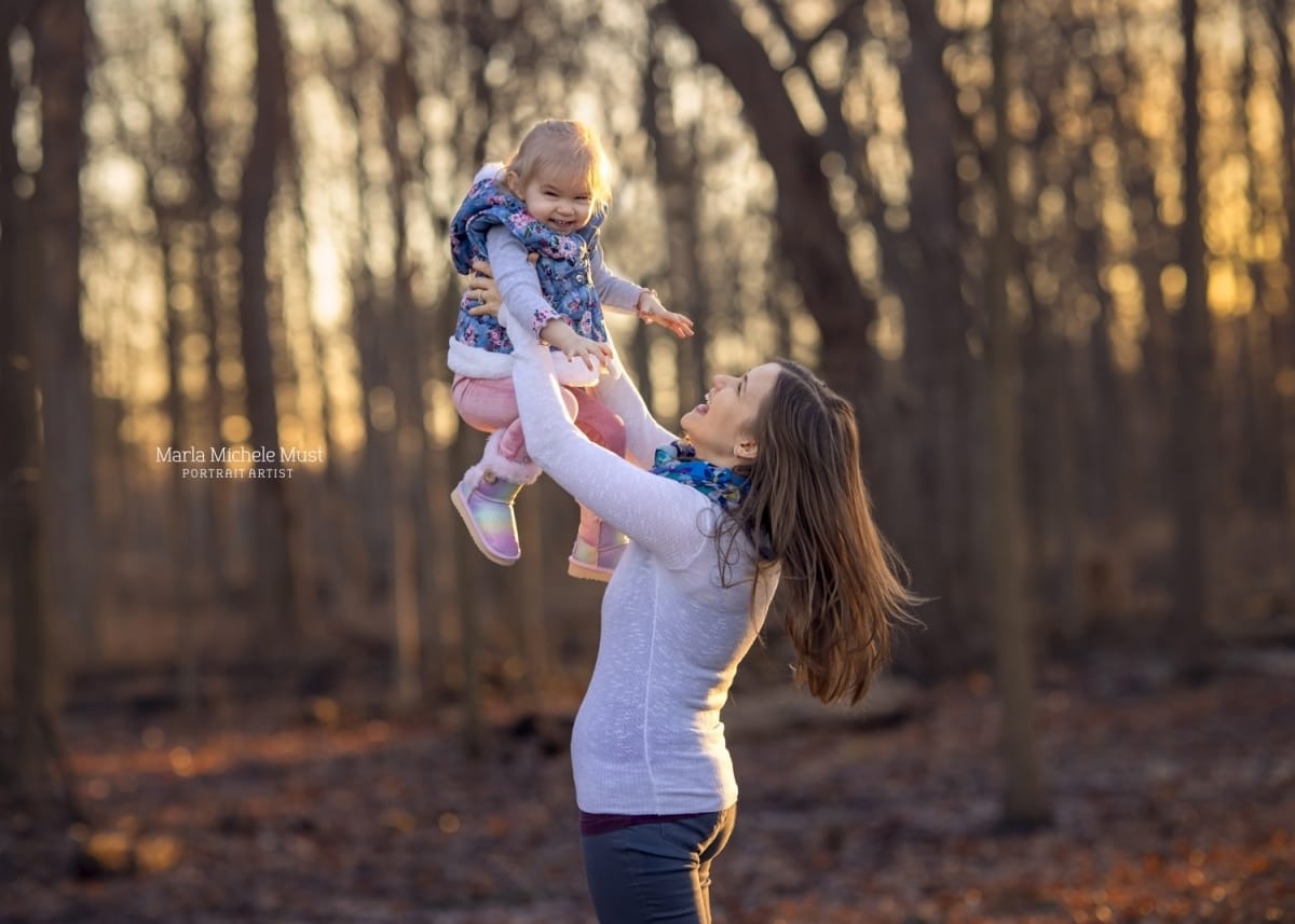 Lovely photo of a Detroit mother and daughter smiling as the mother lifts the toddler in the air, a fall background behind them
