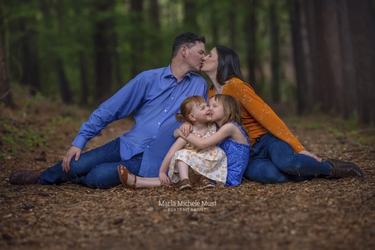 Happy Detroit family photoshoot with a kissing mother and father leaning over their two daughters while sat on a forest floor.