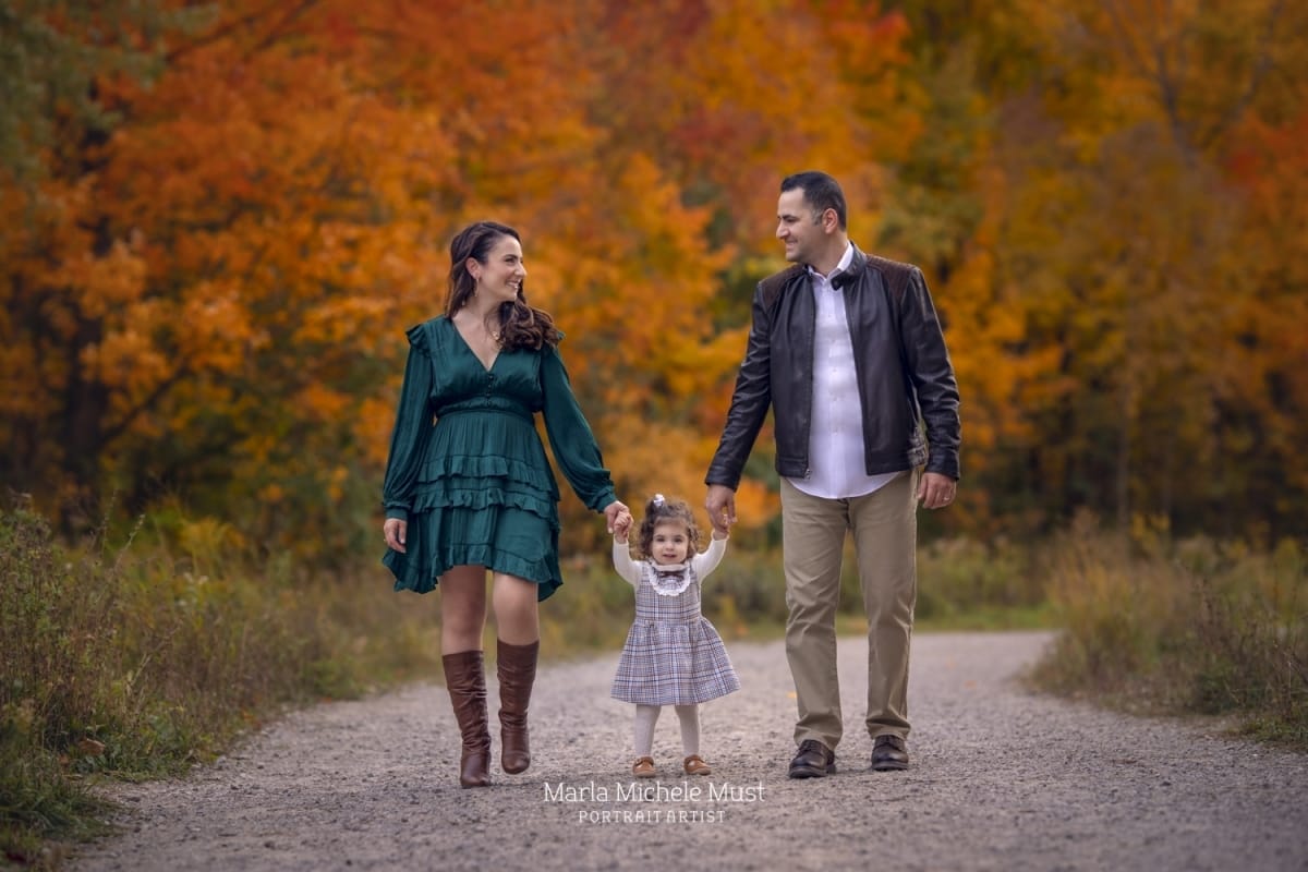 Family portrait of a man and woman walking side by side with their toddler in the middle on an autumn path near Detroit, Michigan