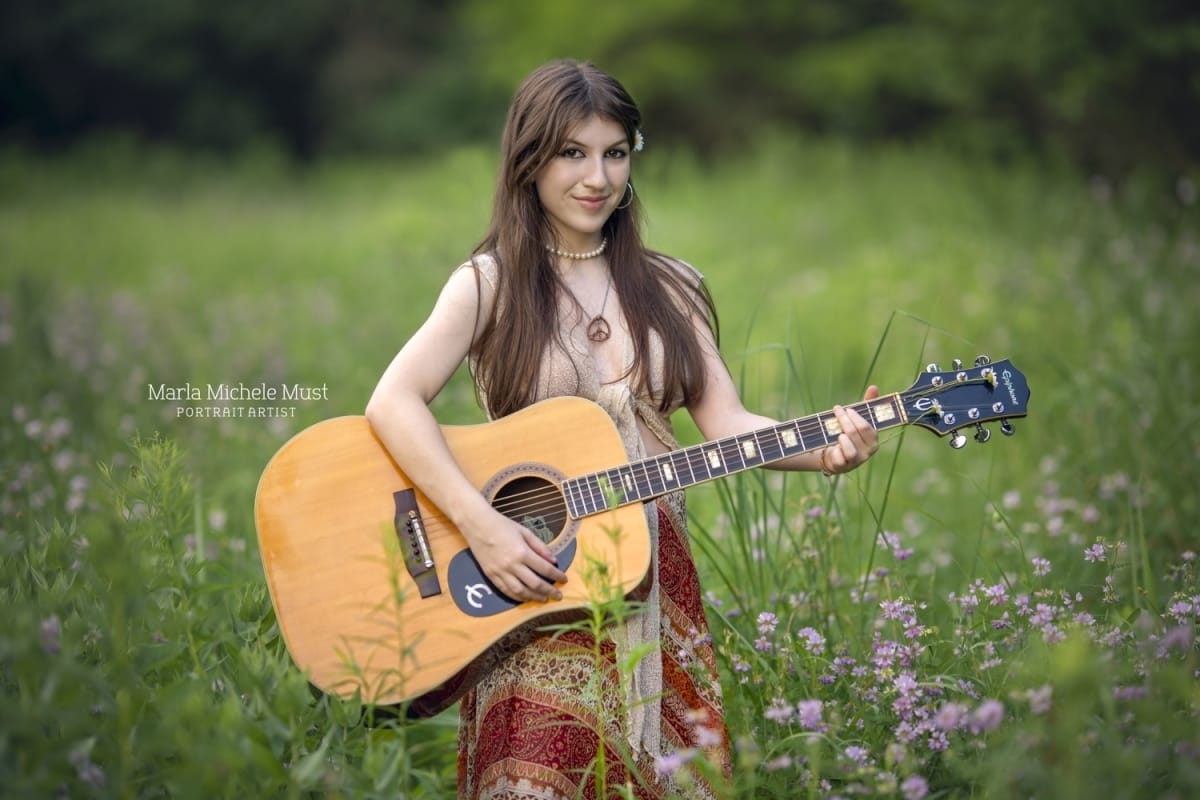 A flower and 70's aesthetic senior photoshoot in a Michigan park as a girl poses while holding her acoustic guitar.