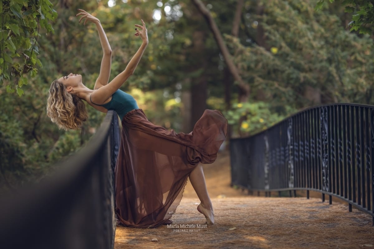 Dancer gracefully embodies a lyrical pose, gracefully extending her arms long while leaning her back against a bridge, captured by a talented Detroit photographer on a Michigan metro area bridge in a forested area.