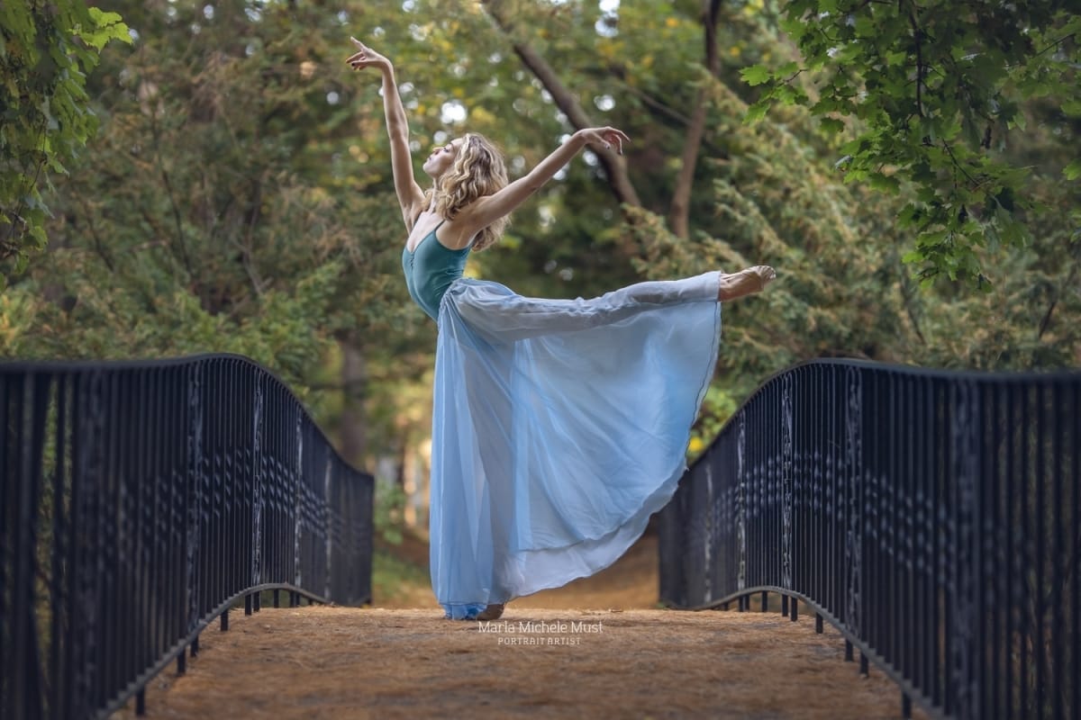 Dancer poses in arabesque while wearing a flowing blue skirt, expertly captured in a Michigan forest during a portrait session by a Detroit photographer.