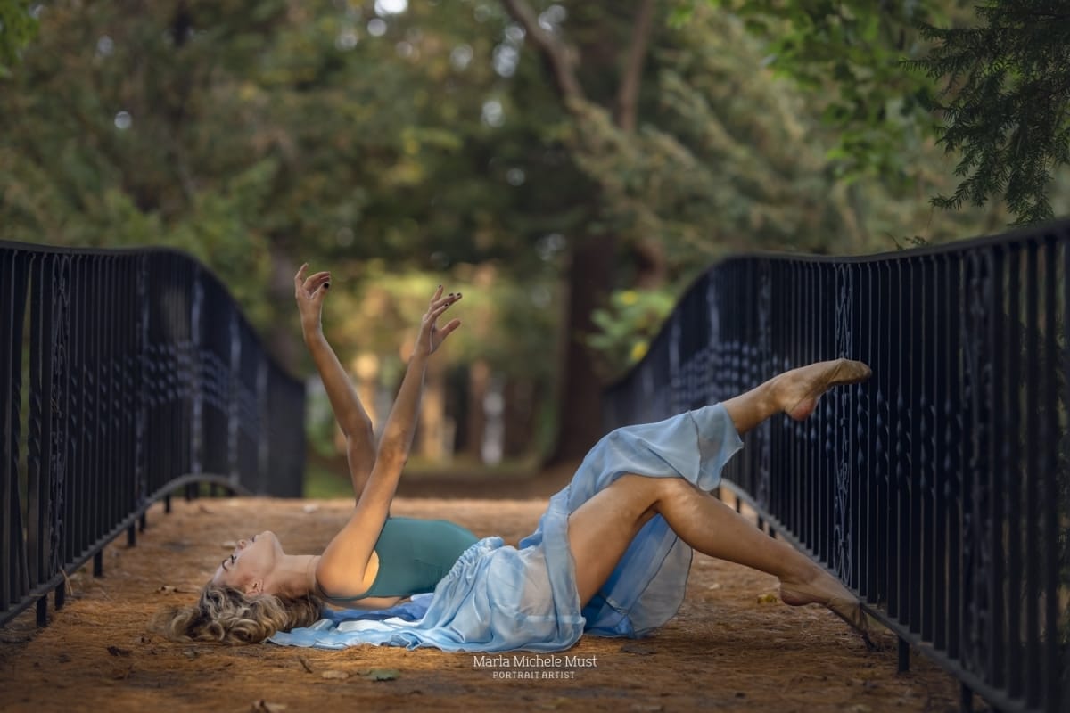 Michigan dancer gracefully strikes a lyrical pose, gracefully extending her arms long while laying on her back in profile, captured by a talented Detroit photographer on a Michigan metro area bridge in a forested area.
