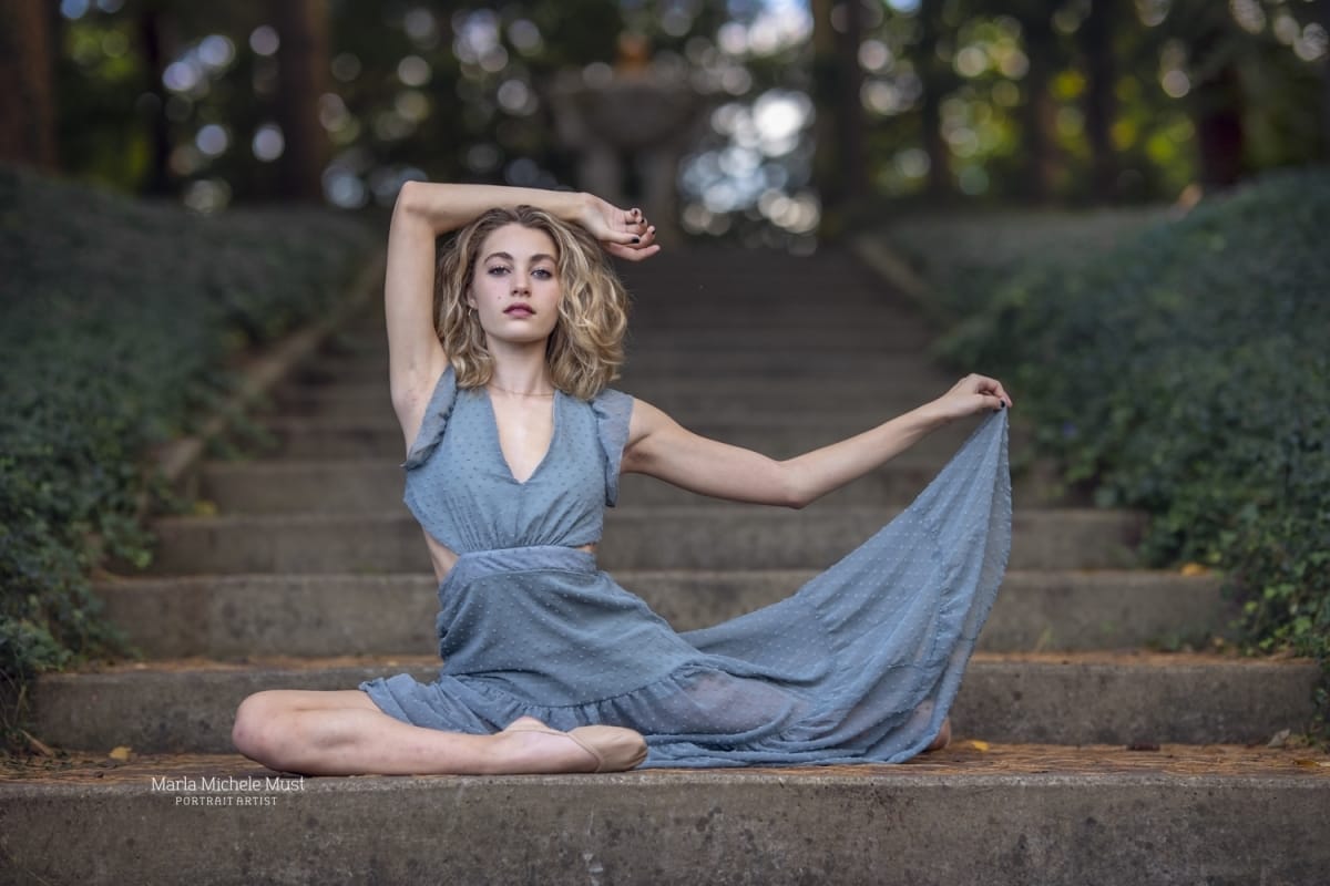 Michigan dancer strikes a powerful pose with seamless flow as she sits on a set of stone steps, one hand holding her dress, the other poised above her head.