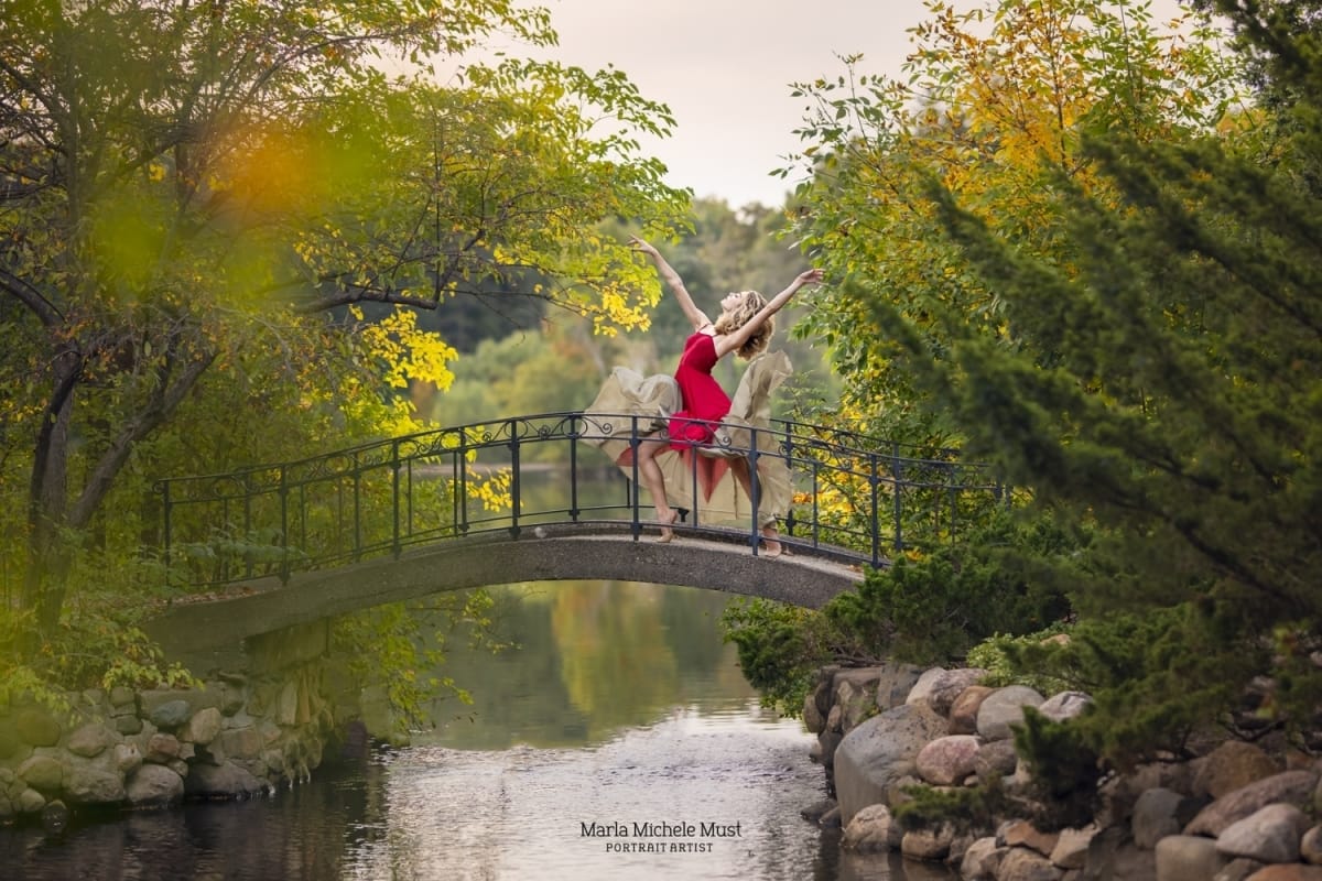 Dancer gracefully poses in a red dress on a bridge, a forest behind her and water flowing below, expertly captured at a distance in a Michigan forest during a portrait session by a Detroit photographer.