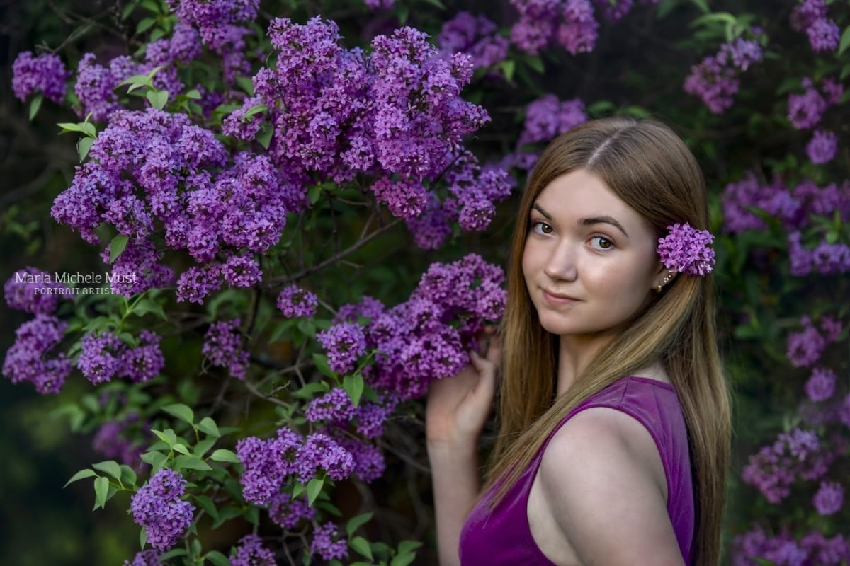 A high school senior's portrait depicting a girl wearing a plum colored dress, a flower in her hair, holding the bloom of a flowering bush behind her during a Michigan-based senior photoshoot session.