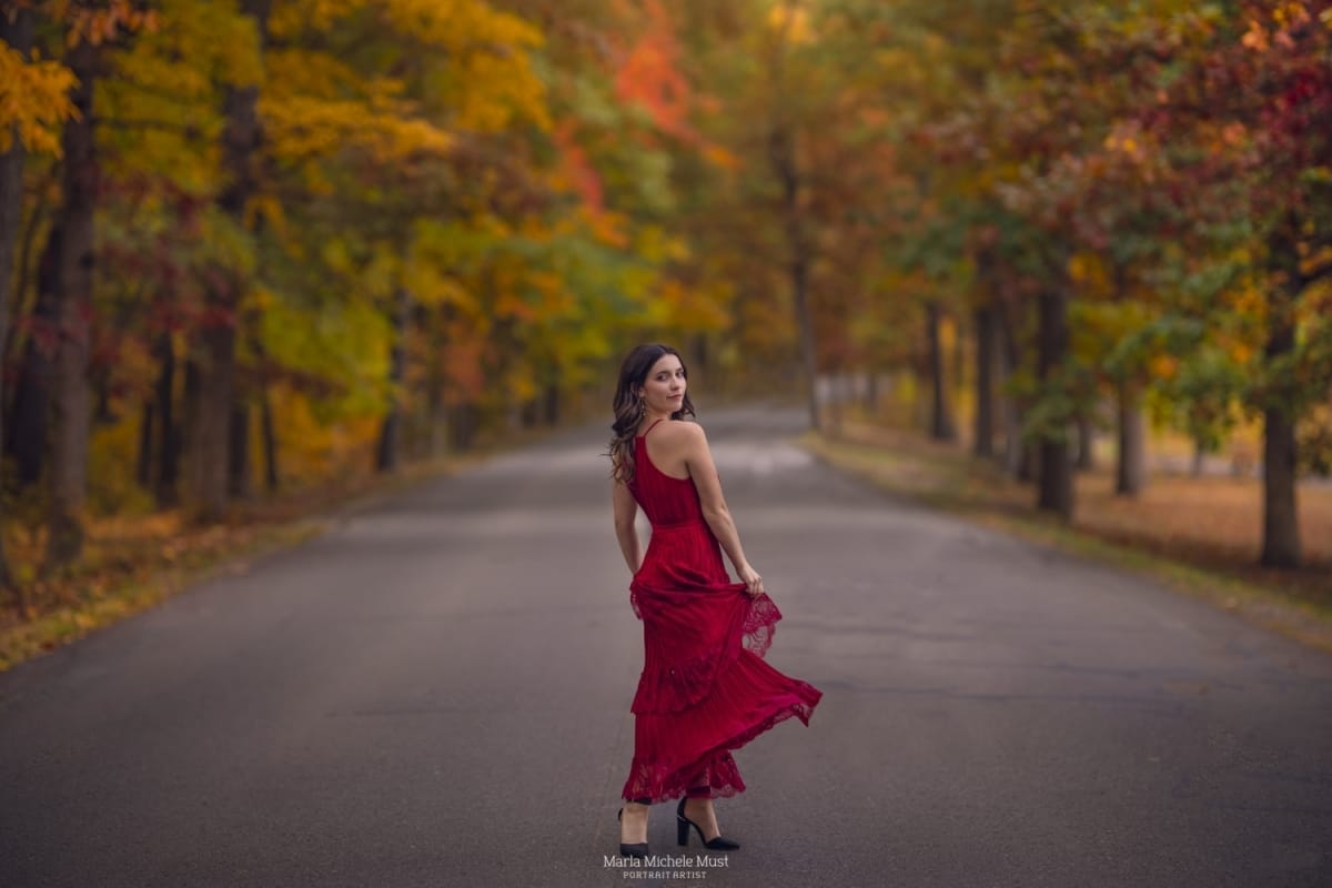 A Michigan high school senior poses for her graduation portrait in a layered red dress with a forest of colorful trees lining the road behind her.