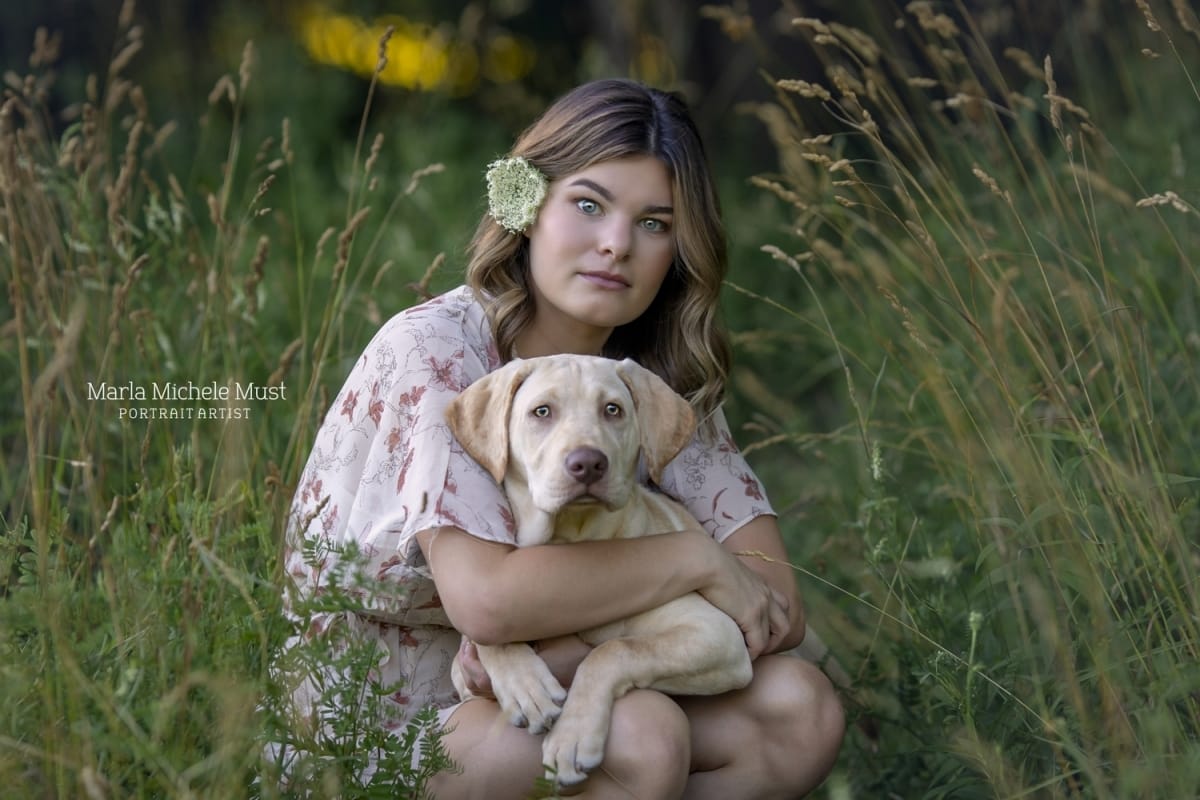 A high school senior holds her golden Labrador retriever for her senior pictures in a flowery Michigan forest