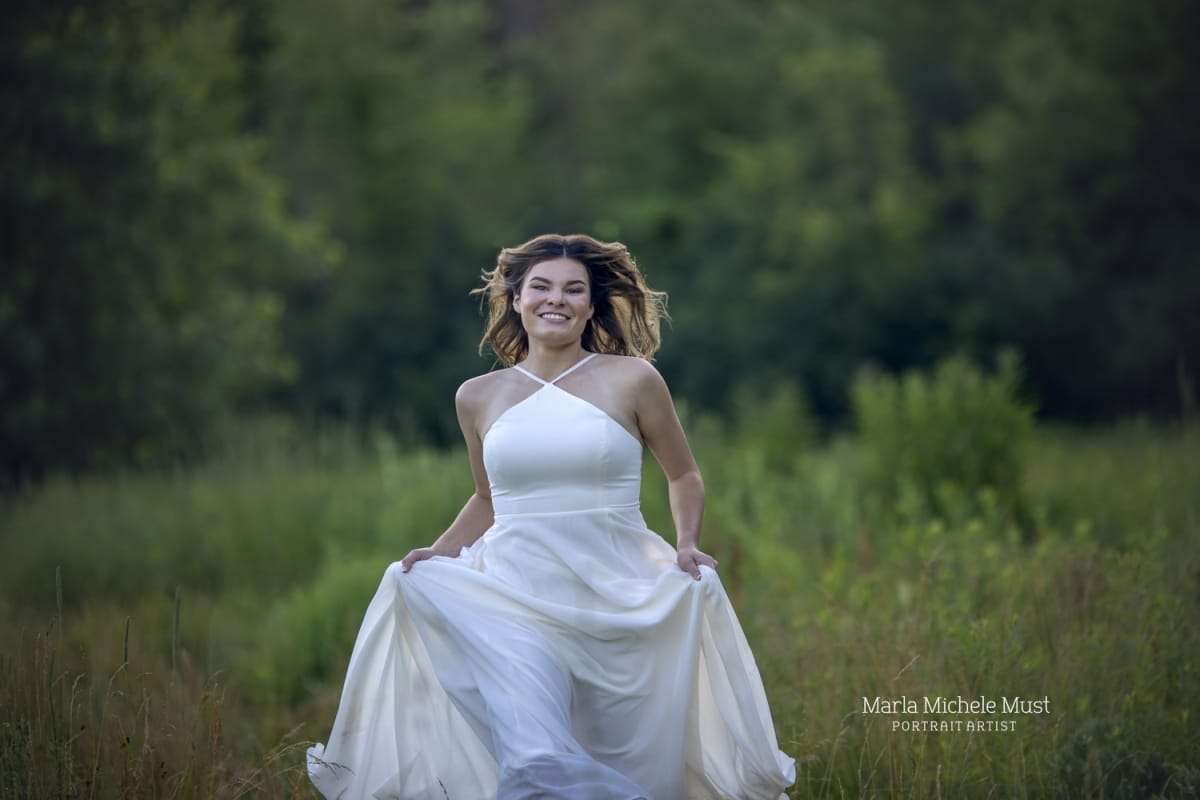 A compelling photo of a high school senior girl in a white dress running through a Michigan Park in a medieval tone, captured during a high school grauduation senior portrait session.