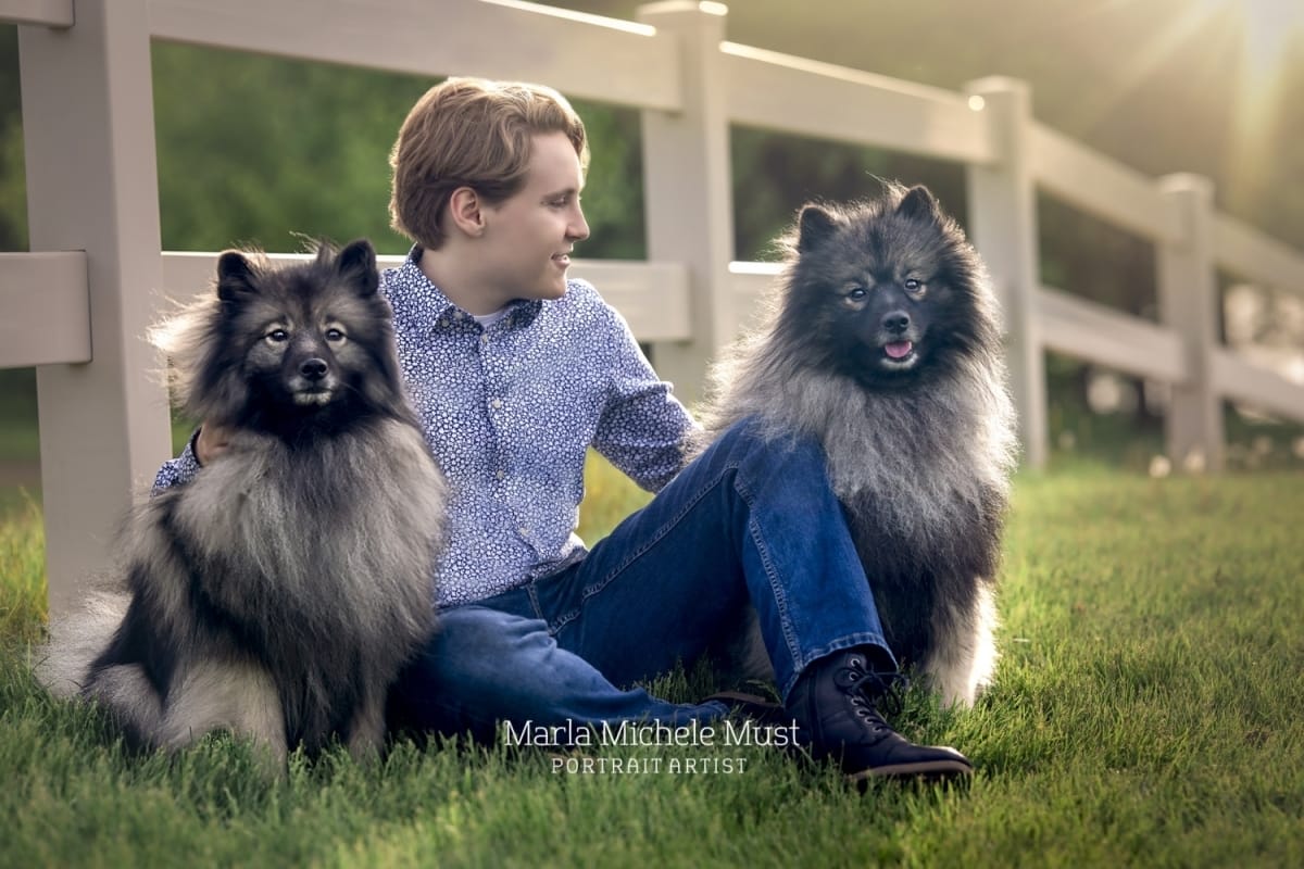 Boy poses for his senior photo with two dogs on either side of them as they sit against a white fence of a Detroit-area farm/ranch.