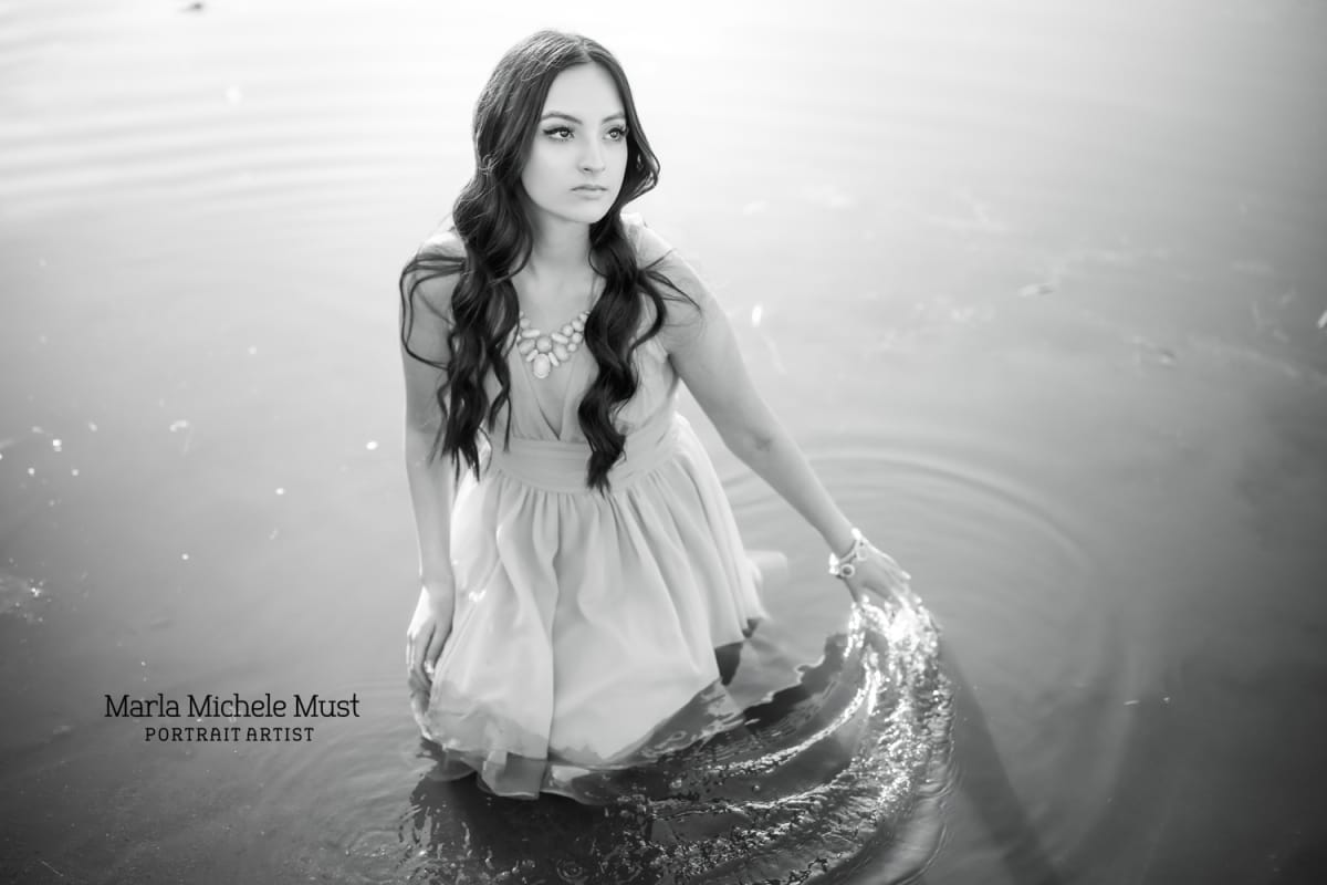 Compelling senior photo of a girl in a floaty whited dress draping her hands along the surface of one of Michigan' great lakes as she wades through during her senior portrait session.