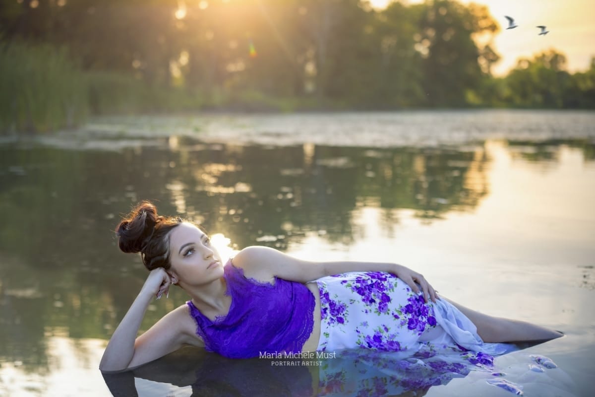 A whimsical, fairy-inspired senior photo of a girl reclining on a beach in a purple dress - one of Michigan's Great Lakes behind her.