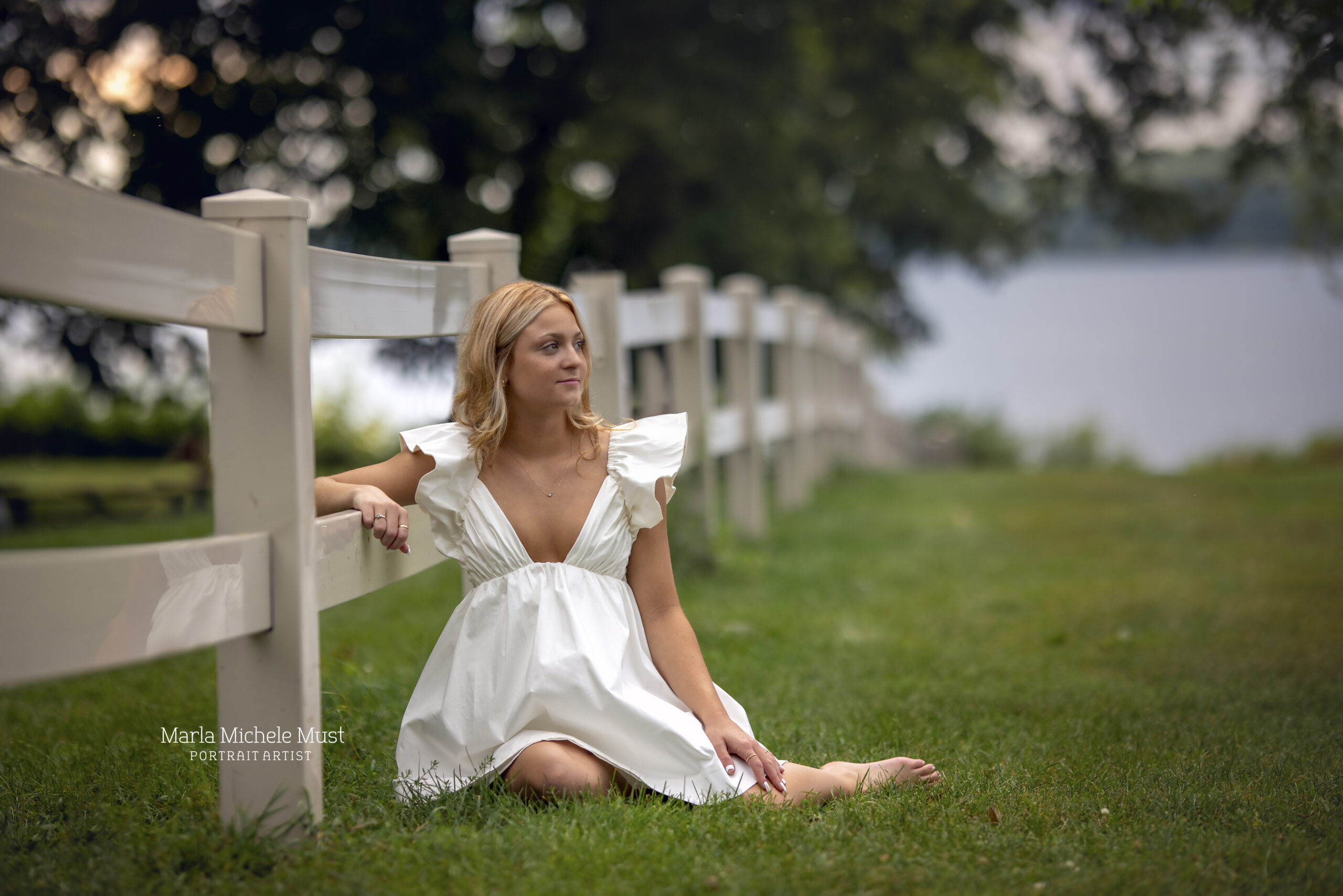 A high school senior stares over her shoulder in a white flowy dress while sitting on the fields of a ranch in Michigan.