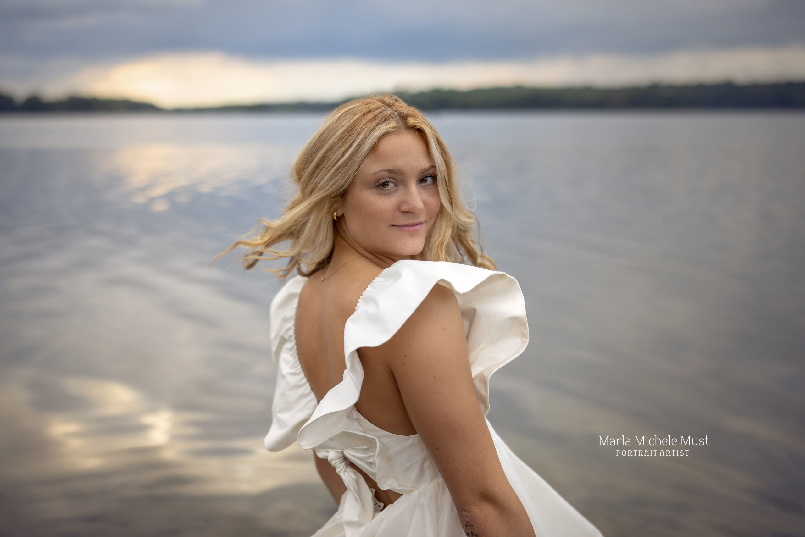 A high school senior stares over her shoulder at the camera in a white flowy dress on the shores of a beach near the Great Lakes in Michigan.