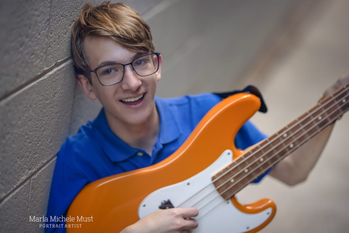 A senior poses with his orange guitar and smiles at the camera - documented by a Detroit photographer during a portrait session