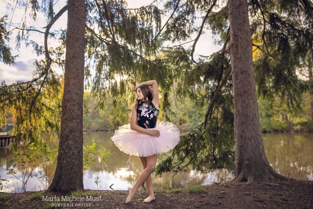 Dancer stands in a Michigan forest, peacefully posing while wearing a pink tutu and black shirt, captured by a Detroit-based photographer during a dance photoshoot.
