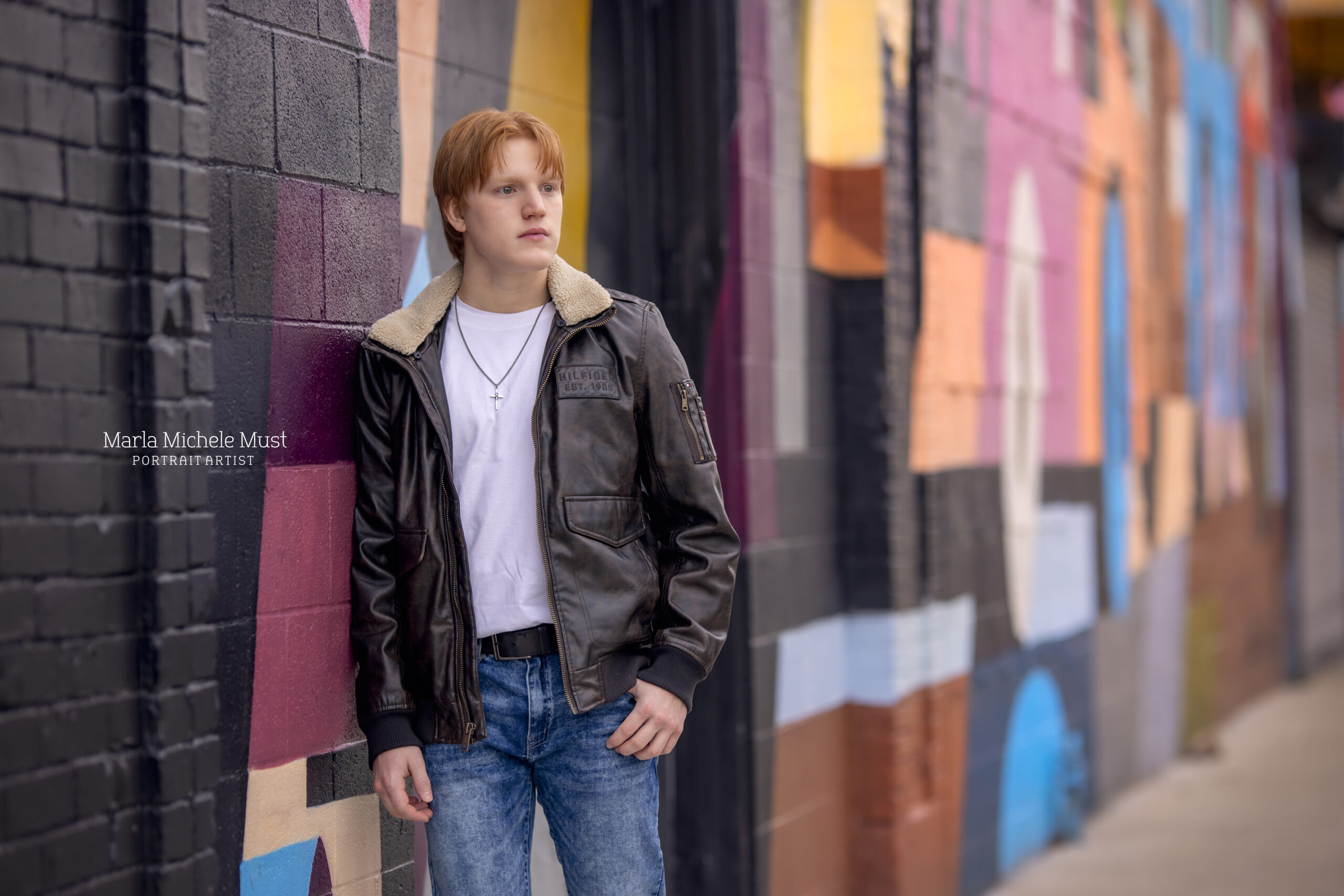 A young man in a leather jacket looks over is shoulder while leaning against a painted wall in Detroit
