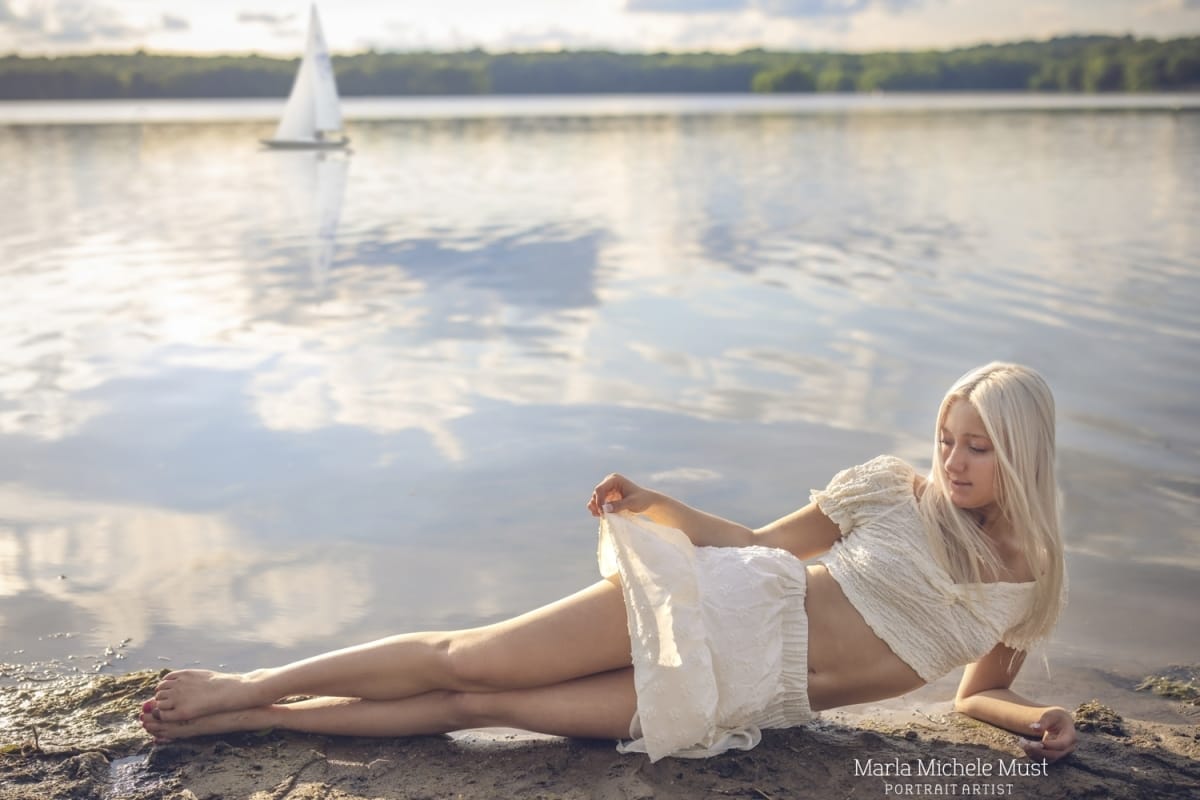 Fairytale inspired high school senior photo of a young woman in a flowing white dress lying in front of a Michigan Great Lake with sailboat in background