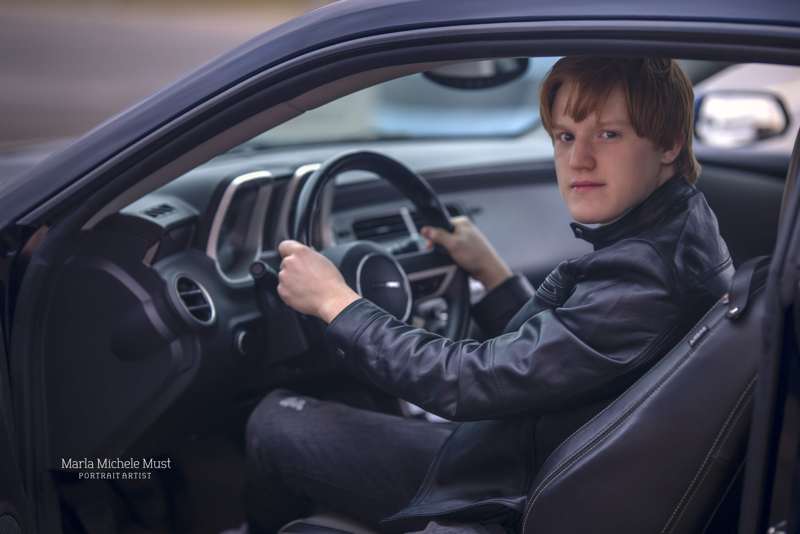 A young man in a leather jacket looks over is shoulder while holding the steering wheel of a sports car in Detroit