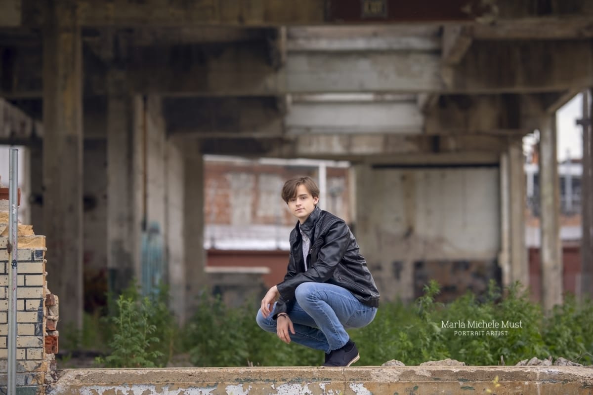 Senior poses in a leather jacket while crouched in a forested urban area in downtown Detroit.