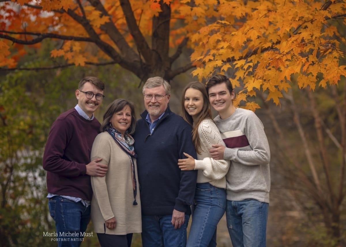 A family portrait during the fall with a couple standing with their two children under a tree with orange leaves.