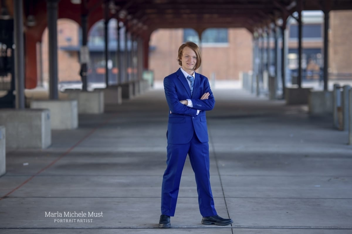 Young man poses for his senior portrait in Downton Detroit while wearing a blue suit, arms crossed