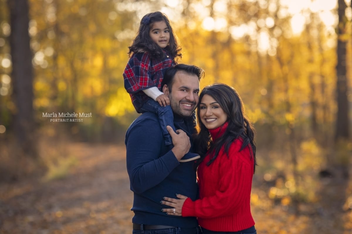 A family portrait of a mother and father wearing blue and red, their young daughter sitting the father's shoulders.
