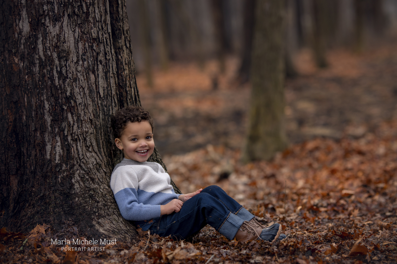 A child is sitting and leaning back against a tree, and smiling to the camera as many autumn leaves surround the forest floor around him near Detroit.