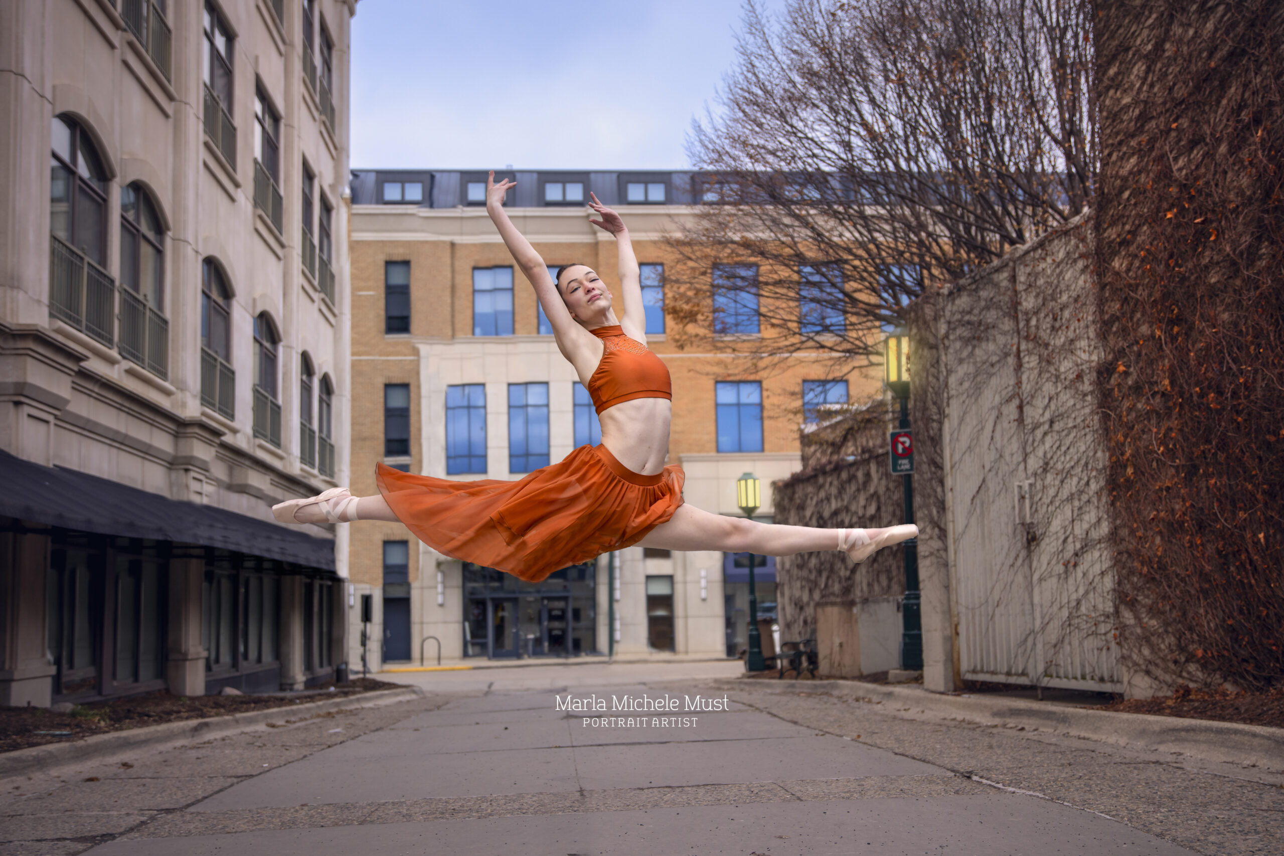 Skilled dancer wearing coordinating orange satin two-piece outfit exhibits ethereal grace with a soaring leap, beautifully photographed by a Detroit-based portrait photographer.
