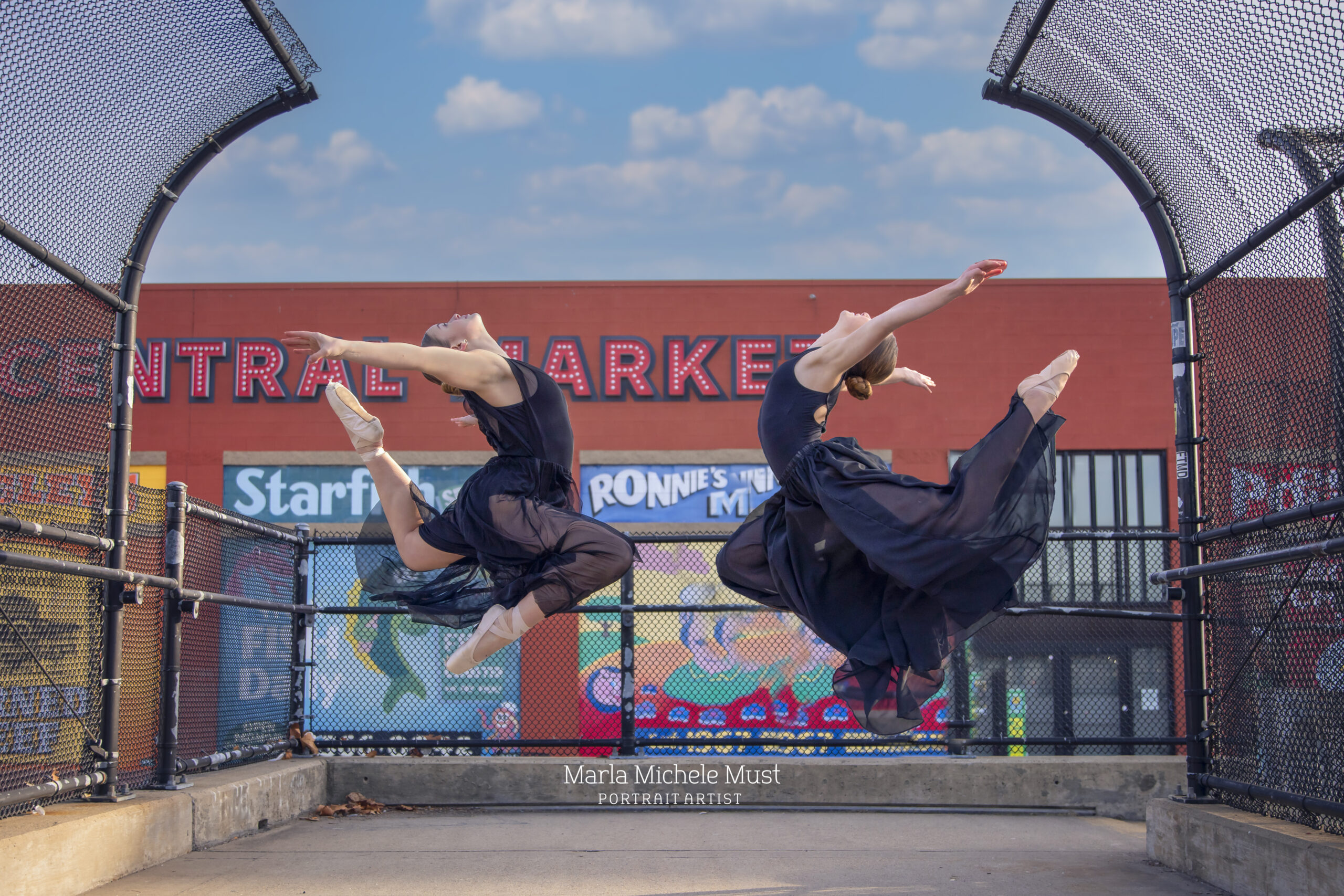 Two Detroit-area dancers mirror each other in identical black, flowing dresses and poses gracefully executed while soaring in the air with legs and arms extended behind them.