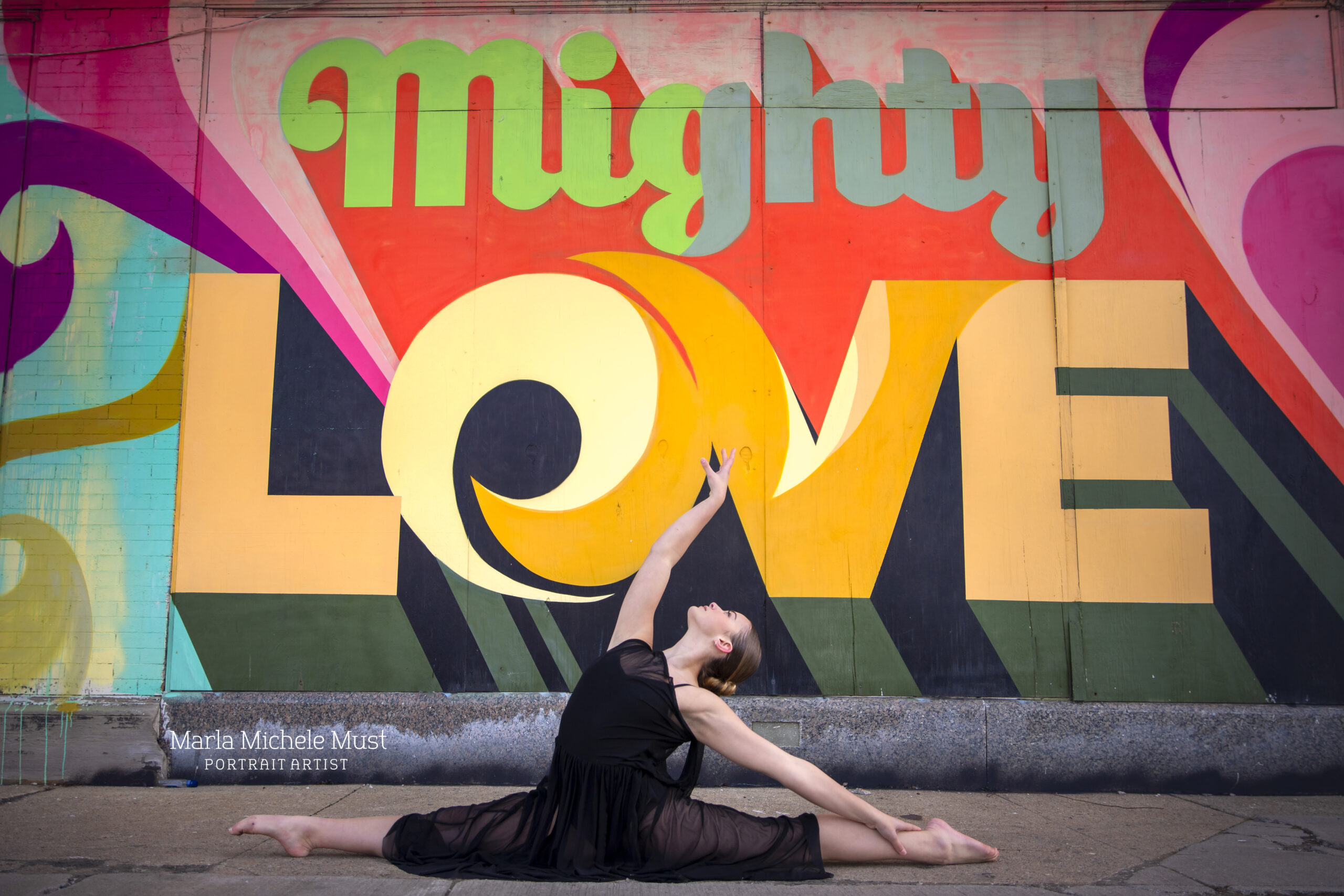 Posed against a bright mural stating the phrase Mighty Love, a dancer does the splits in a flowing black dress, her arm gracefully extended to the sky..