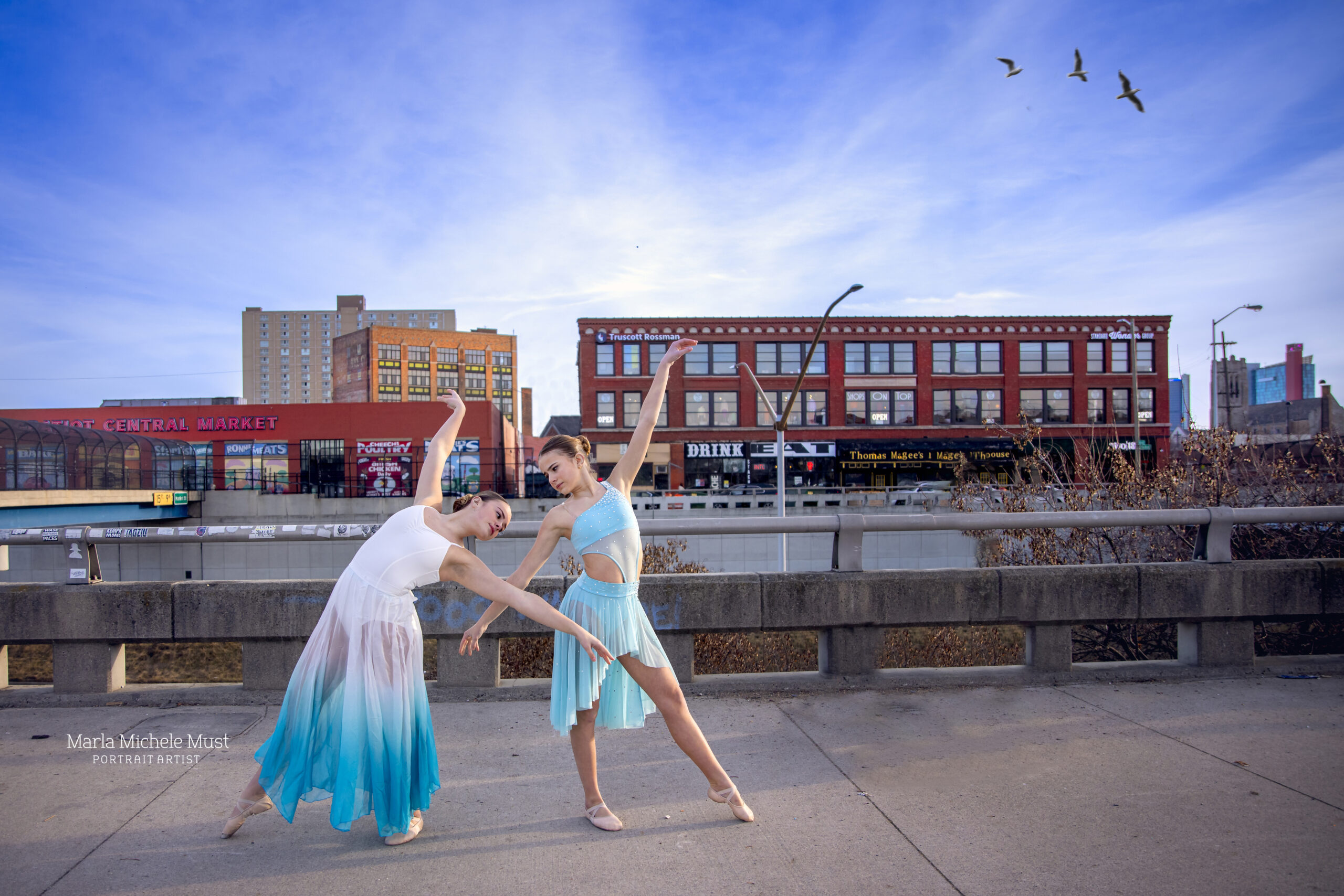 Two dancers gracefully embody a lyrical pose while wearing matching blue dresses among a Michigan metro area cityscape during a Detroit photoshoot with a skilled dance photographer.