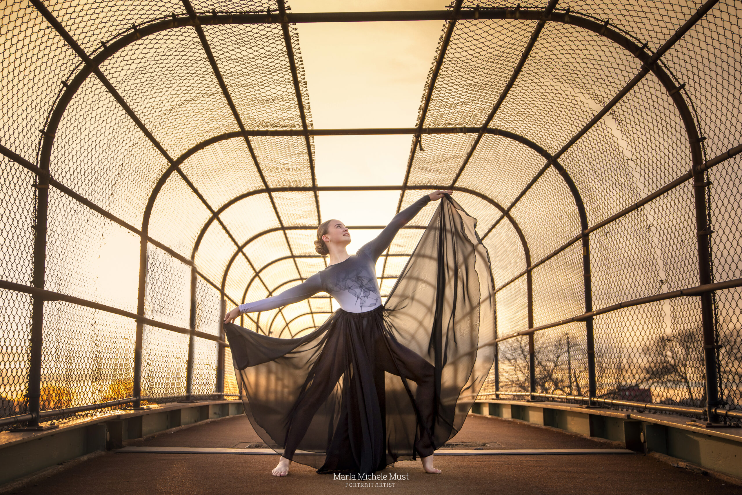At sunset, a dancer holds ends of her flowing black skirt against the sunset behind her during a dancer photoshoot in Detroit.