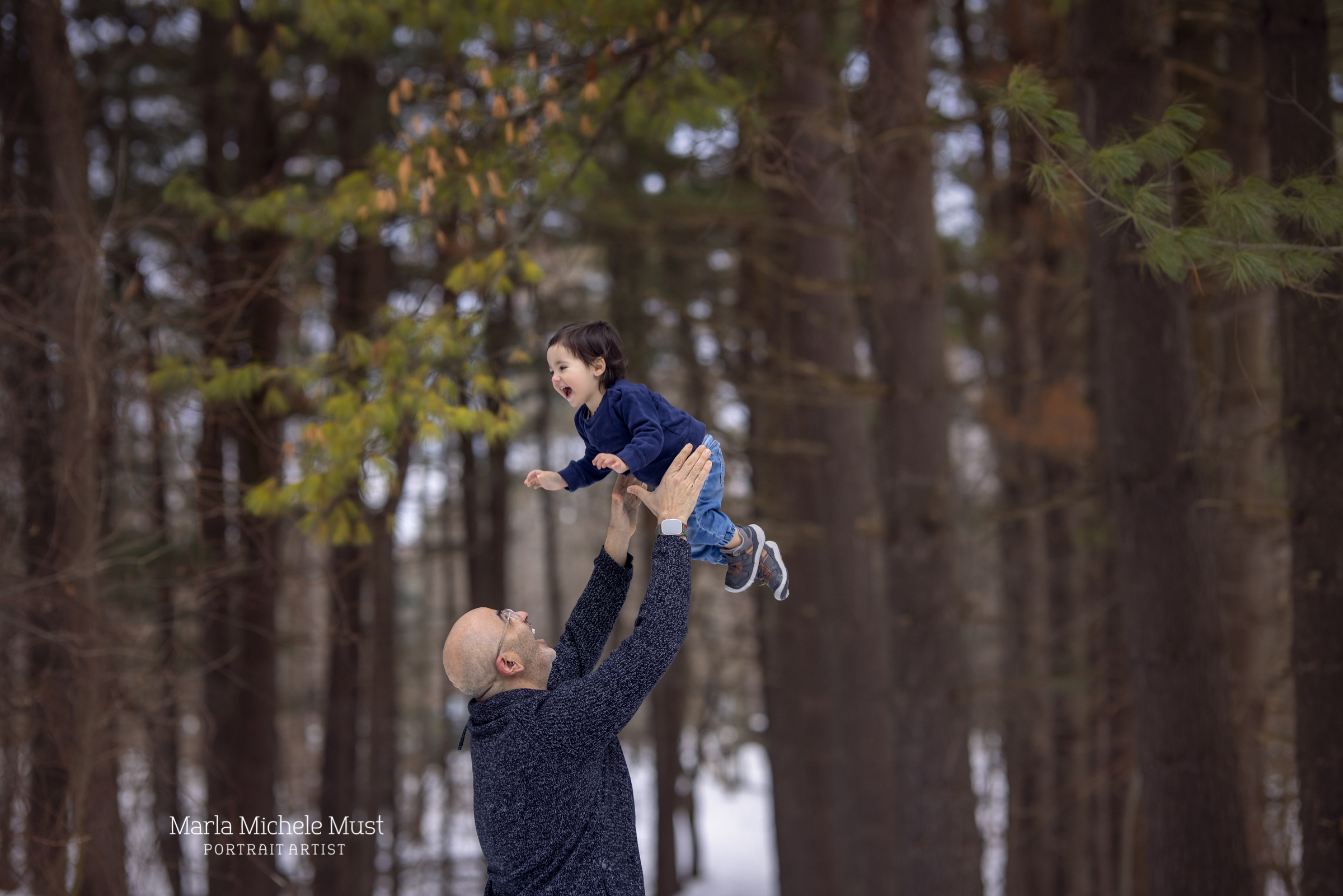 Local family photographer takes photo of aDetroit mother and daughter smiling as the father lifts the toddler in the air, a fall background behind them