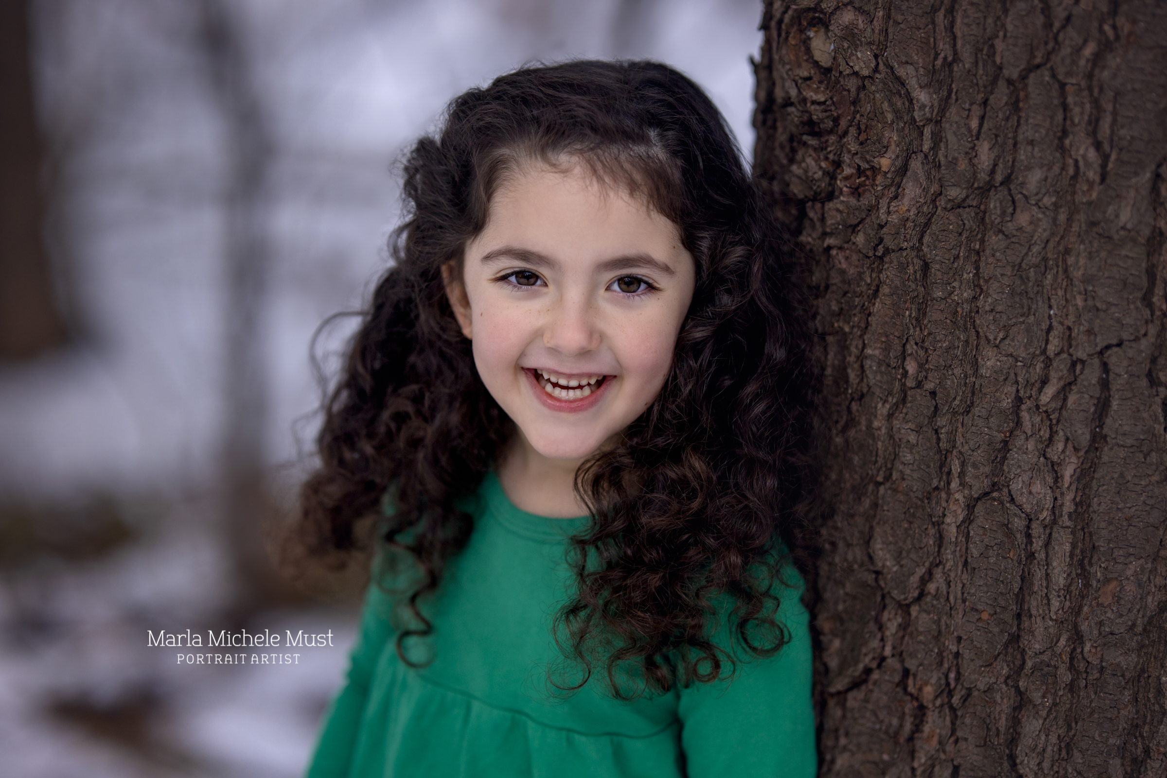 A young girl poses for a family portrait in Detroit. She stands against a snow outdoor background and smiles to the camera. She has brown, curly hair and is wearing a green shirt.