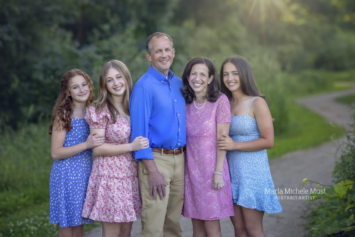 A bright and happy family portrait of a mother, father, and three daughters wearing blue and pink dresses in the countryside.