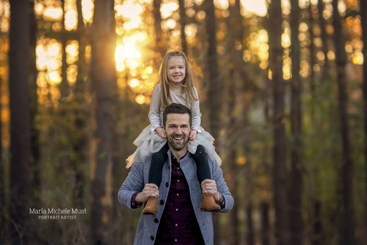 Detroit family photoshoot with daughter sitting on father's shoulders while smiling at the camera in the forest.