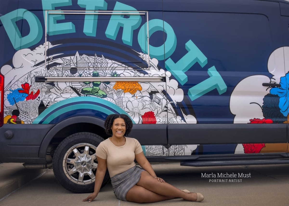 A young graduate sits gracefully in front of a car in front of a painted van stating "Detroit" as part of a high school senior photoshoot