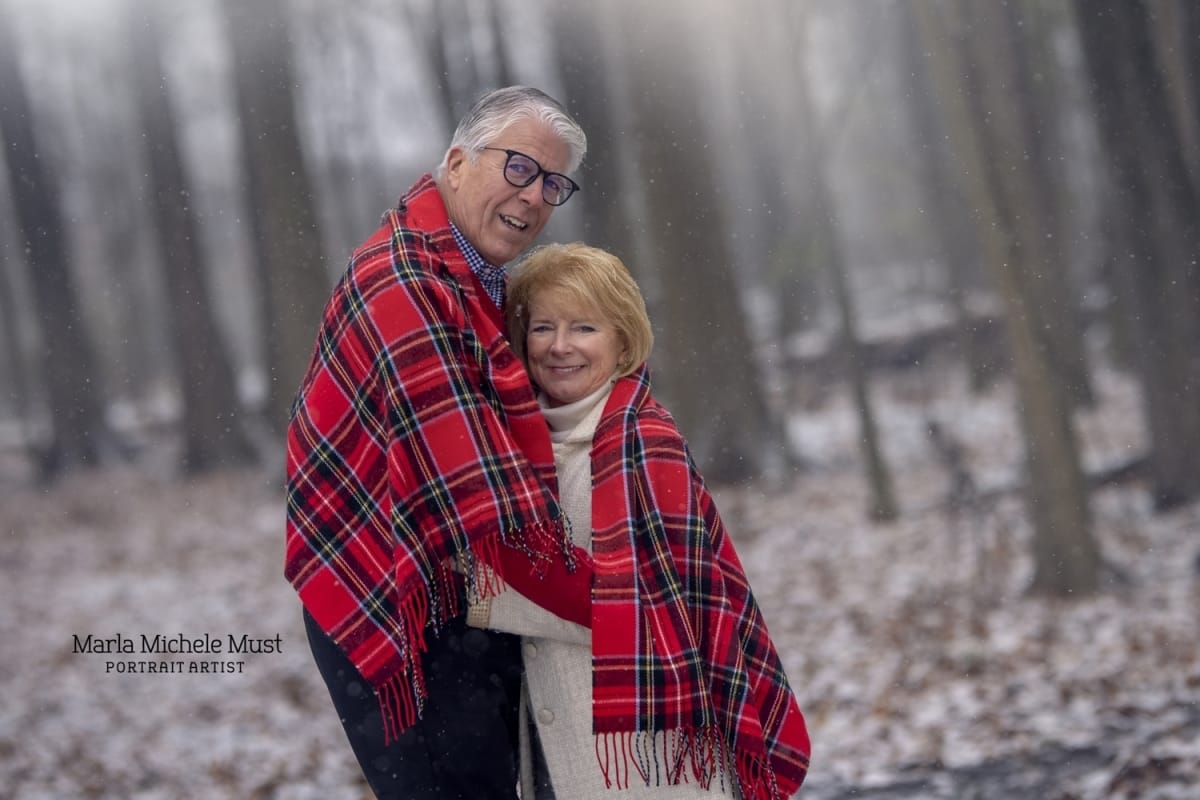 couple smiling at the camera and standing under a red plaid blanket in a snowy forest for a photoshoot near Grand Rapids