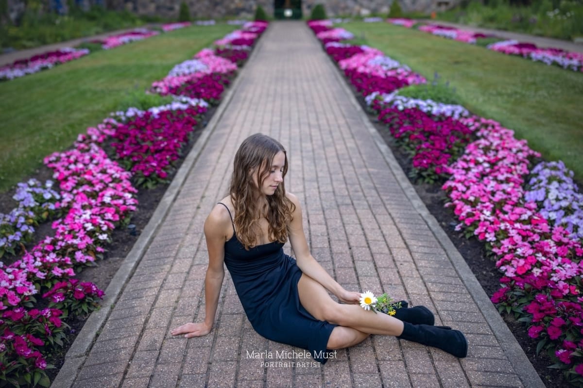 High school graduation photoshoot of a girl in a dress sitting on a lane bordered by flowers